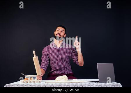 young man baker cooks bread, pasta, pizza looking at laptop sitting at table in studio on black background copyspace Stock Photo