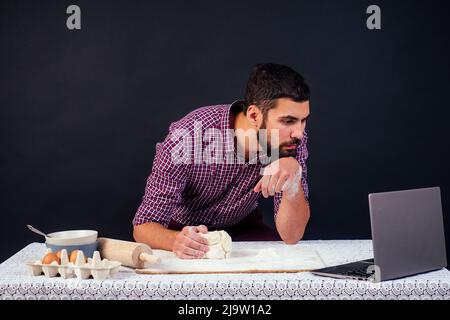 young man baker cooks bread, pasta, pizza looking at laptop sitting at table in studio on black background copyspace Stock Photo