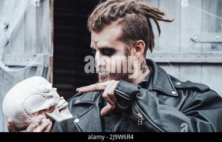 Scary man with dreadlocks looking at skull. Celebrating halloween. Terrifying black white face monster makeup and stylish costume, image.Horror,fun at Stock Photo