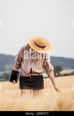 Farmer inspecting and examining agricultural field before harvest. Woman with straw hat and digital tablet standing in barley field Stock Photo