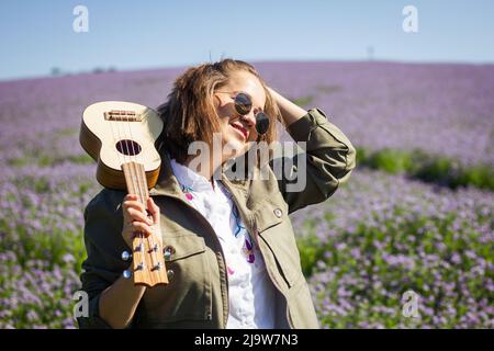 Portrait of happy young smiling woman with ukulele enjoying sunlight in flowering meadow at spring Stock Photo