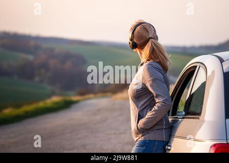Woman with headphones leaning at car and resting during a road trip. Female person enjoying solo travel Stock Photo