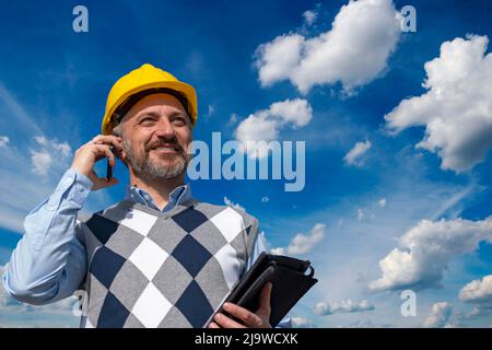 Happy Manager In Yellow Hardhat Standing And Using Smart Phone Outdoors. Stock Photo