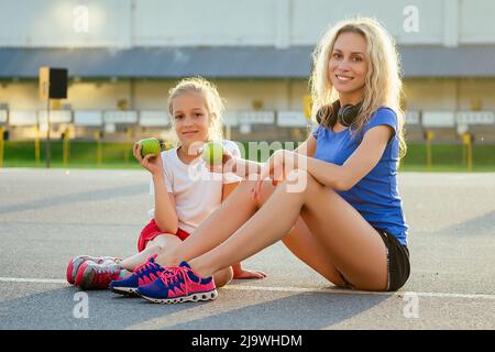 young beautiful blonde female person with a little girl in sportswear sitting on the asphalt and eat a green apple at the stadium. Active family Stock Photo