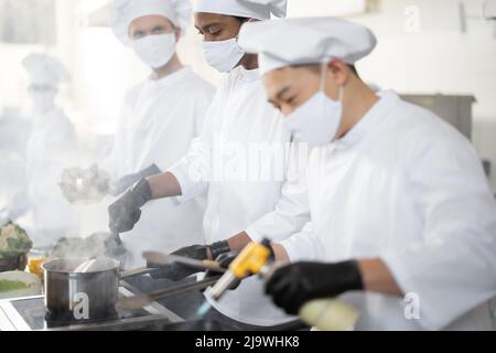 Multiracial team of chef cooks in white uniform cooking together in the kitchen. Asian chef frying meat with burner, latin and european guy cooking on background. Cooks wearing face masks and protective gloves  Stock Photo