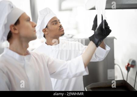Two well-dressed chefs look on printed checks with orders while cooking in the professional kitchen. Latin-American and caucasian guys working together at restaurant Stock Photo