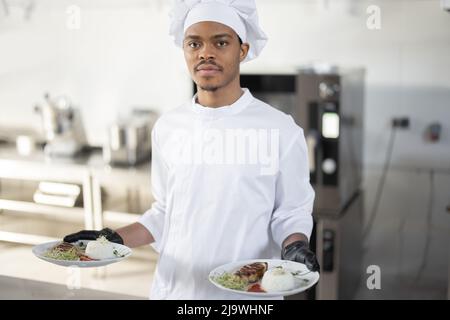 Portrait of young handsome smiling chef in uniform standing with ready meal in the kitchen. Happy Latin-American cook working at restaurant. Concept of professional occupation and haute cuisine Stock Photo