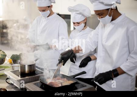 Multiracial team of cooks in uniform and face masks cooking meals for a restaurant in the kitchen. Concept of teamwork at restaurant during pandemic. Latin, Asian and European guys cooking together Stock Photo