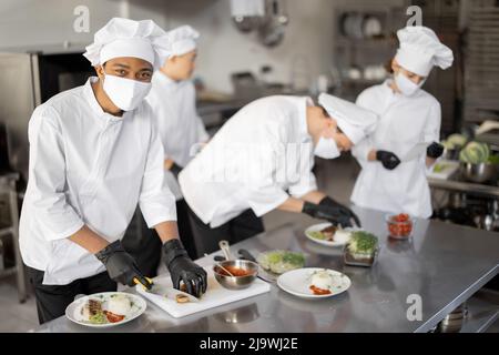 Multiracial group of cooks finishing main courses while working together in the kitchen. Cooks wearing uniform and face mask. Team prepares meals for the restaurant Stock Photo