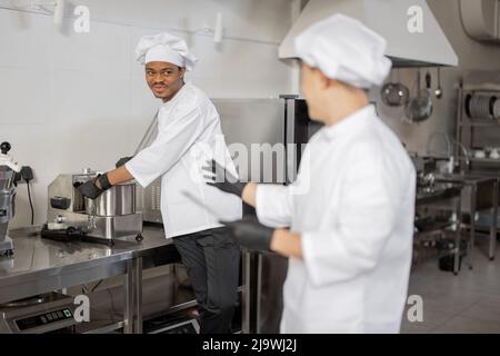 Two smiling chefs cooking in restaurant kitchen. Asian chef tells latin guy what dish to cook while reading the order. Concept of teamwork at restaurant Stock Photo