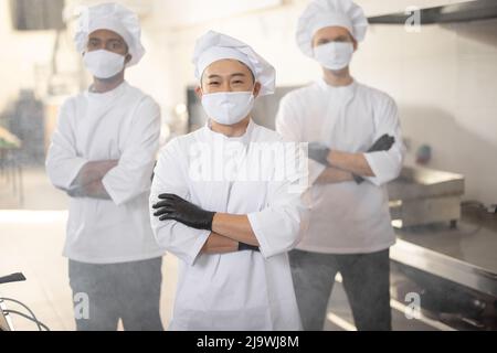 Portrait of three well-dressed chef cooks with different ethnicities standing together in restaurant kitchen. Asian chef standing in front, latin and european guys on background. Cooks wearing face masks ready to cook during pandemic Stock Photo