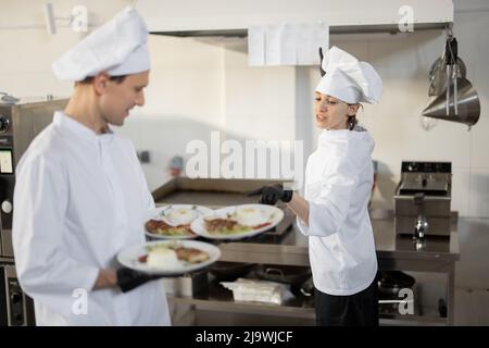 Chef standing with ready meals, talking with female cook in the kitchen. Happy caucasian cook working at restaurant. Concept of professional occupation and teamwork Stock Photo