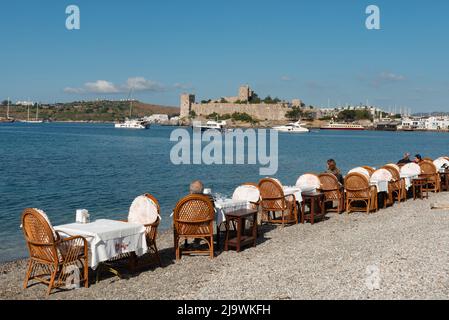 Restaurant Tables on the beach in Bodrum harbour with Bodrum Castle in the background, the Aegean coast of south west Turkey. Stock Photo