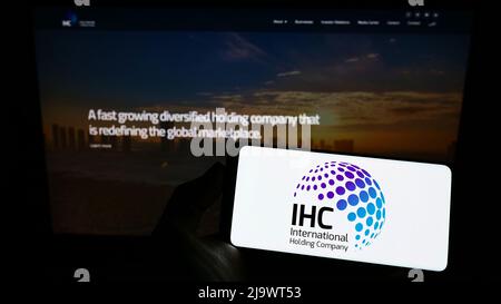 Person holding smartphone with logo of Emirati International Holding Company PJSC (IHC) on screen in front of website. Focus on phone display. Stock Photo