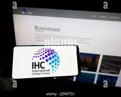 Person holding mobile phone with logo of Emirati International Holding Company PJSC (IHC) on screen in front of web page. Focus on phone display. Stock Photo
