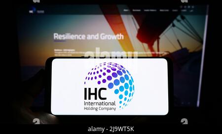 Person holding cellphone with logo of Emirati International Holding Company PJSC (IHC) on screen in front of webpage. Focus on phone display. Stock Photo