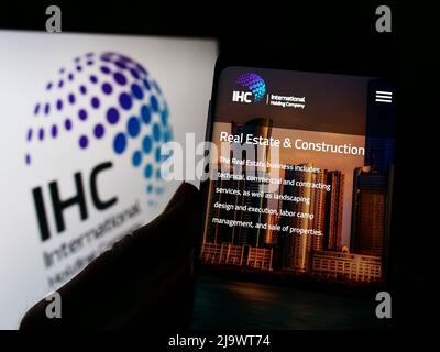 Person holding smartphone with webpage of Emirati International Holding Company PJSC (IHC) on screen with logo. Focus on center of phone display. Stock Photo