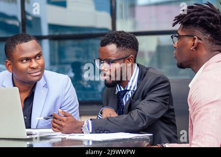 A group of three stylish African American students entrepreneurs in fashion business suits working sitting at table with laptop in a summer cafe Stock Photo