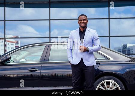 successful businessman handsome African American man in a stylish suit in a blue jacket standing in front of a cool new black car on the street Stock Photo