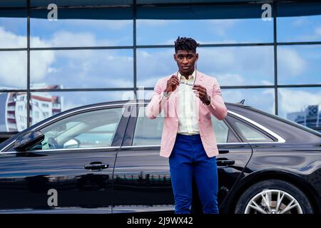 successful businessman handsome African American dreadlocks man in a stylish suit in pink jacket standing in front of a cool new black car on the Stock Photo