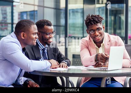 A group of three stylish African American students entrepreneurs in fashion business suits working sitting at table with laptop in a summer cafe Stock Photo