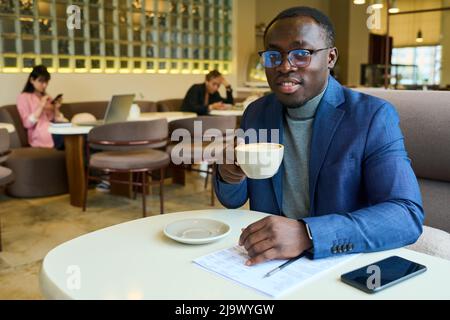 Portrait of African businessman in eyeglasses smiling at camera while sitting at table in cafe and drinking coffee Stock Photo