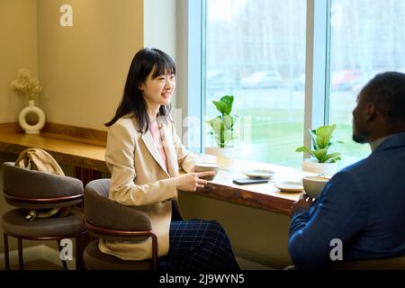 Asian young businesswoman drinking coffee and talking to customer during their meeting at cafe Stock Photo