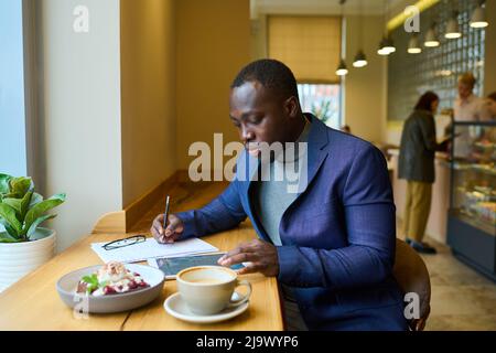 African serious businessman using tablet pc and filling document at table in cafe during his breakfast Stock Photo
