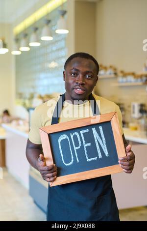 Portrait of African young waiter in uniform holding open sign and looking at camera, he opening new coffee shop Stock Photo