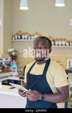 Portrait of African young waiter in uniform smiling at camera while standing in coffee shop and using tablet pc Stock Photo