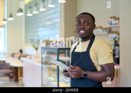 Portrait of African young waiter in uniform smiling at camera while taking order online on tablet pc Stock Photo