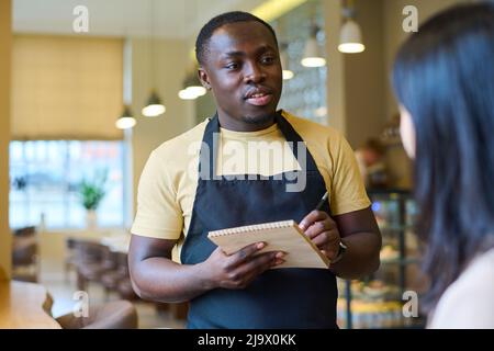 African American waiter making notes in notepad while talking to woman in cafe, he taking order from her Stock Photo