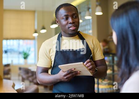 African American waiter making notes in notepad while talking to woman in cafe, he taking order from her Stock Photo