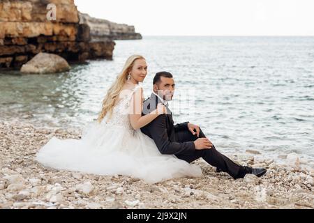 Portrait of young blonde bride in white dress and brunet groom in suit embracing and sitting on rocky sea coast in Italy side view, seascape Stock Photo