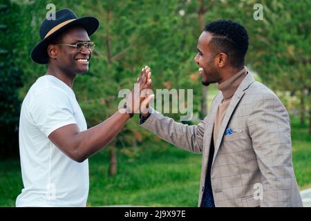 two black men in stylish suits meeting in a summer park. African-Americans friends hispanic businessman embrace high five greeting each other teamwork Stock Photo