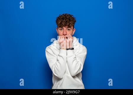 Young caucasian man with curly hair isolated for blue background dressed in a white sweatshirt, covering his mouth with his hands and round eyes Stock Photo