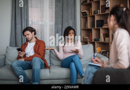 Sad young black wife shows at caucasian hasband with smartphone on consultation with psychiatrist Stock Photo