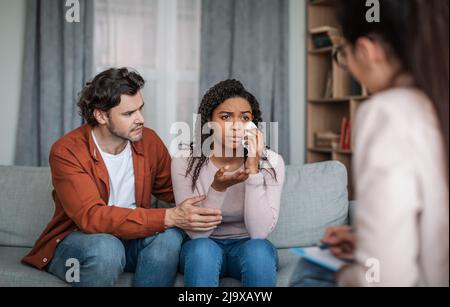 Sad young european male calm crying black female, talk to psychiatrist in clinic interior Stock Photo