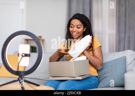 Unboxing video. Excited black female blogger recording content for her vlog, unpacking parcel with new shoes at webcam Stock Photo