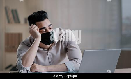 Bored tired hispanic guy worker in medical mask during pandemic coronavirus young business man tired ill worker feel boredom in home office hold head Stock Photo