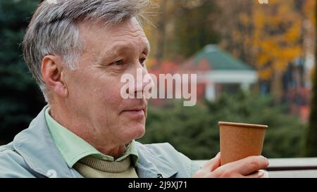 Dreamy calm gray-haired elderly pensioner man grandfather middle aged adult male person sitting on autumn park bench drinking coffee hot tea Stock Photo