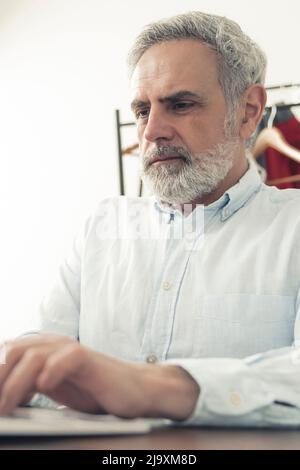 Vertical indoor shot of a focused elderly man with gray hair, wearing baby blue shirt and using keyboard. Work concept. High quality photo Stock Photo