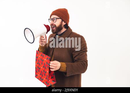 Caucasian man with a megaphone. Studio shot of a millennial bearded guy in a beanie and brown coat holding a loudspeaker and a gift bag over white background,. High quality photo Stock Photo