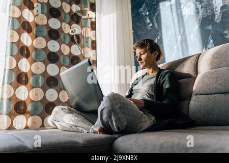 A woman sits comfortably on the couch, legs crossed, and uses a laptop Stock Photo