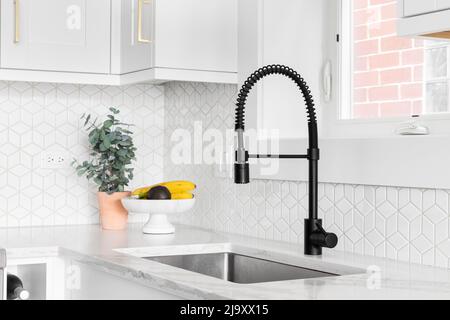 A farmhouse kitchen sink detail shot with a black faucet, mosaic tile backsplash, marble countertops, and white cabinets. Stock Photo