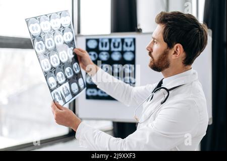 Experienced smart focused doctor looking at x-ray at his cabinet, caucasian male therapist in white medical uniform, with stethoscope concentrated learning patient's x-ray, standing in medical office Stock Photo