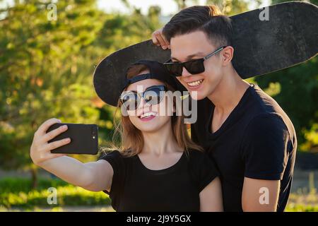 active couple in love hugging in the summer park. beautiful woman in a cap and sunglasses is holding a skateboard(longboard) and photographed take Stock Photo