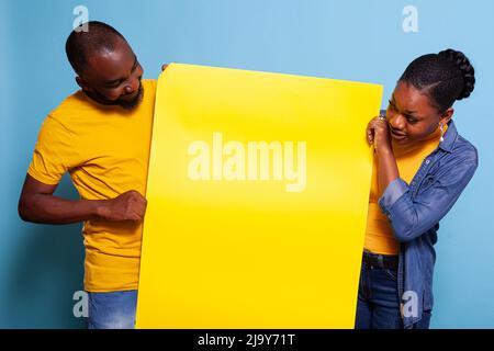 Young partners holding copy space placard for advertisement. People showing blank cardboard banner to write message and pass information. Cheerful couple with mockup poster in studio. Stock Photo