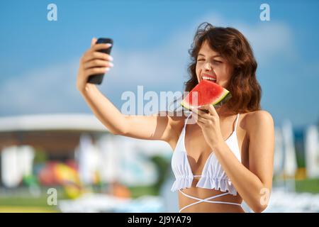 Pretty lady in bikini taking selfie, while biting tasty slice of watermelon on vacation. Portrait view of cute girl eating watermelon, holding sell phone to take funny photo at day. Concept of selfie. Stock Photo