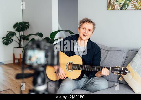 Young man playing acoustic guitar and recording music video for his social networks. Amatuer musician create content at home by modern camera. Musicia Stock Photo